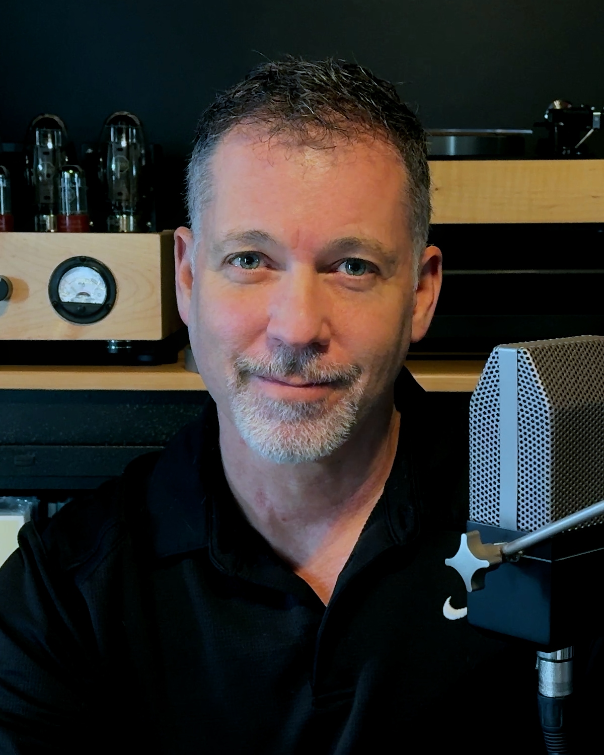 Rick Coste in his listening room, with a tube amplifier, a turntable and a studio microphone behind and beside him.
